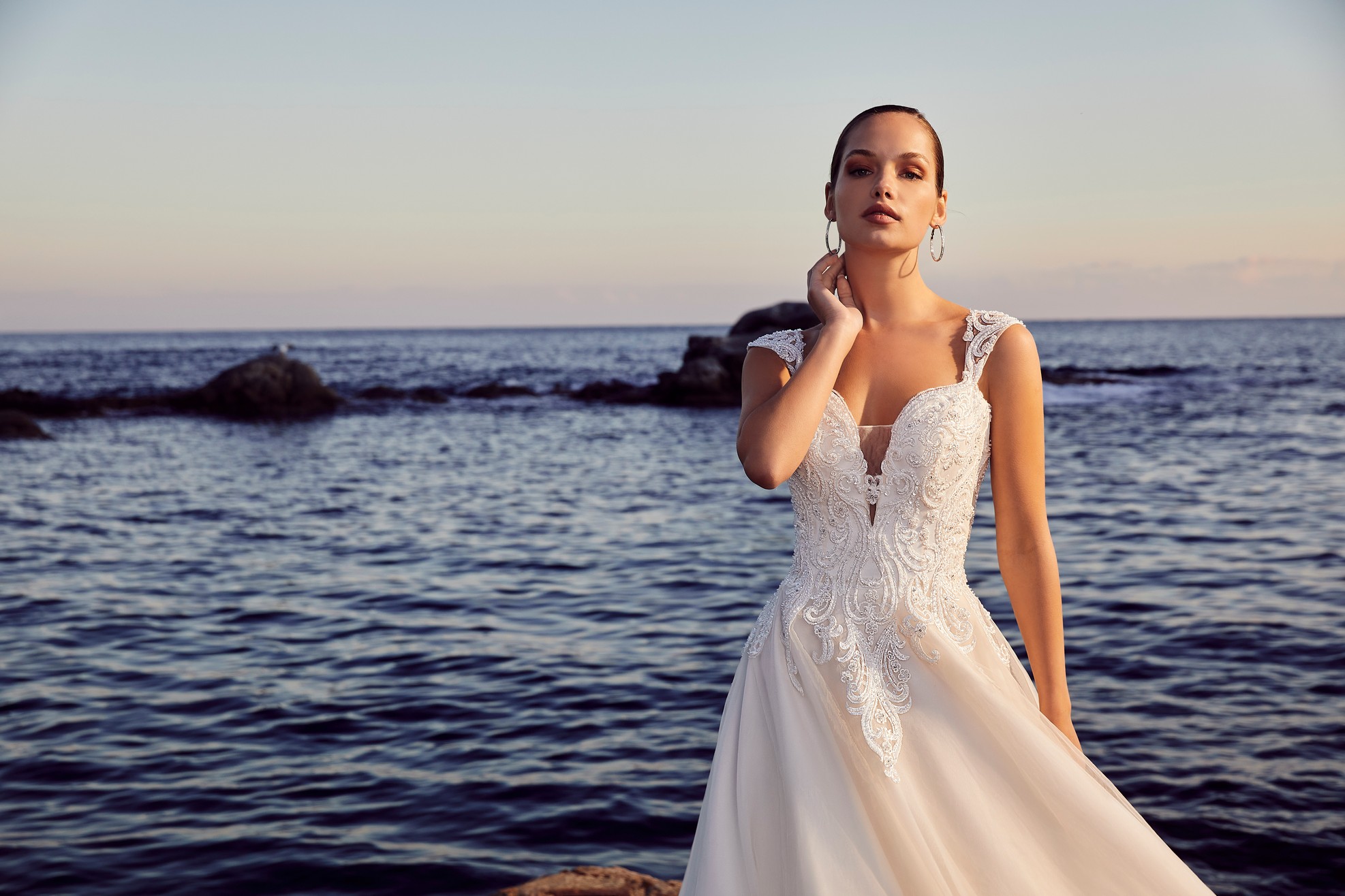 Lady standing on beach next to the sea wearing ballgown wedding dress with lace straps and plunging illusion neckline detail
