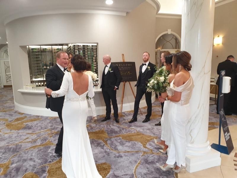 Real bride Emma stood with husband Brian inside a posh entrance lobby. Emma has her back to the camera showing the open back and bustle of Ronald Joyce 18202 wedding dress