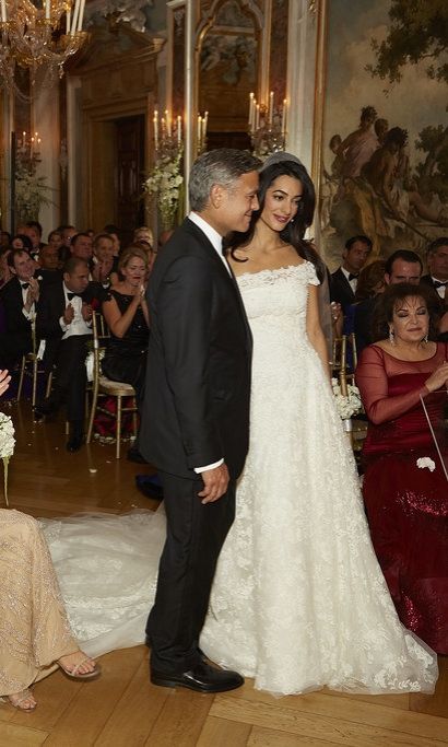 George and Amal Clooney on wedding day wearing lace fit and flare with off the shoulder neckline and cathedral veil