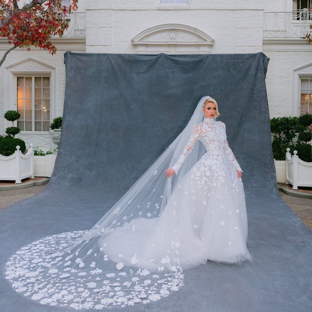 Paris Hilton poses in front of grey phot backdrop on wedding day wearing high neck floral detail ballgown wedding dress