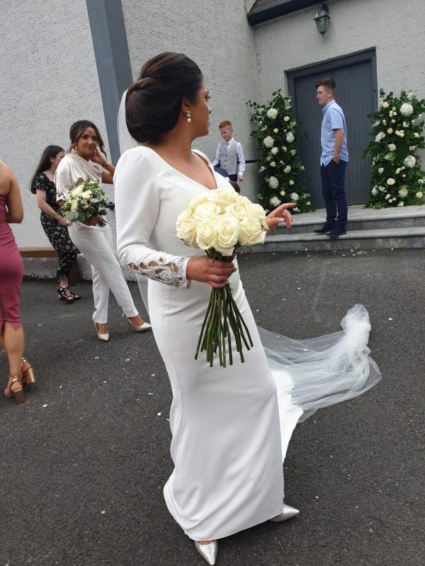 Real bride Emma stood outside near guests holding a bunch of white roses in Ronald Joyce 18202, a fitted sheath wedding dress with long lace cuff sleeves and a v neckline.
