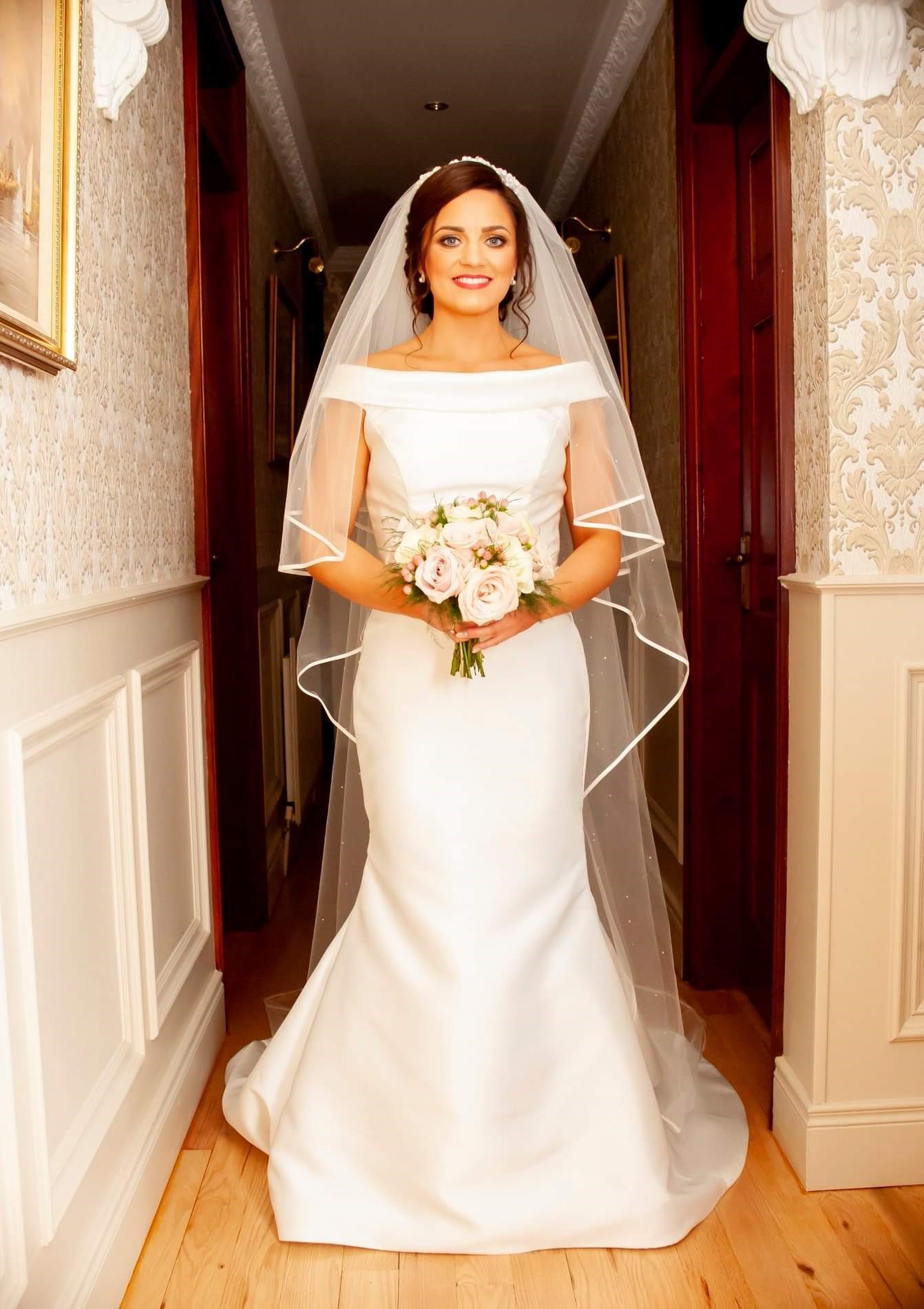 Real bride Sinead stood in a hallway in Ronald Joyce 69153, a plain ivory Mikado fit and flare wedding dress with an off the shoulder bateau neckline and ivory beaded waistband