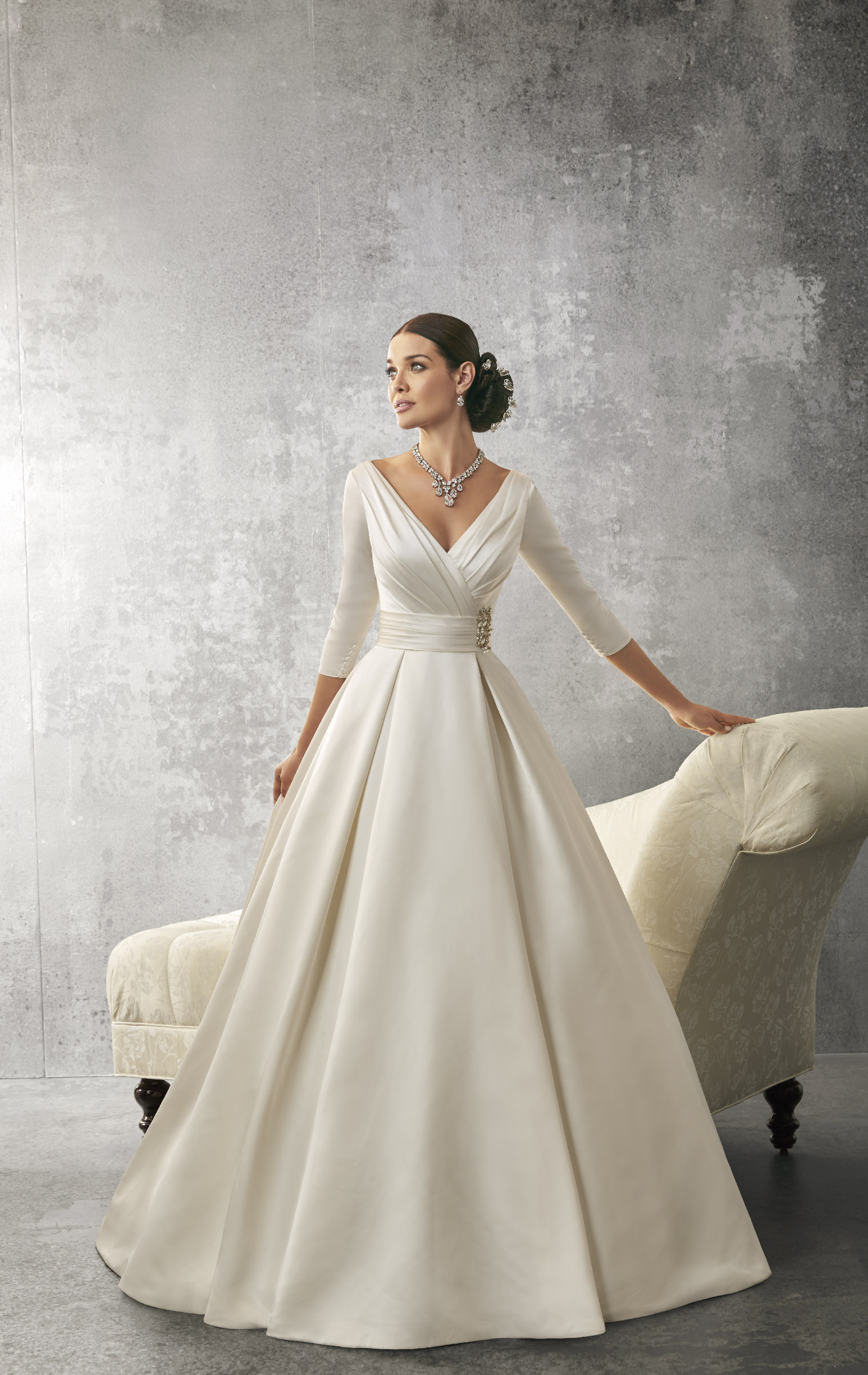 Brunette model stood in front of a cream chaise lounge in Ronald Joyce 69155, an ivory satin ballgown with ¾ sleeves, a v-neckline, and a ruched bodice and waistband with brooch