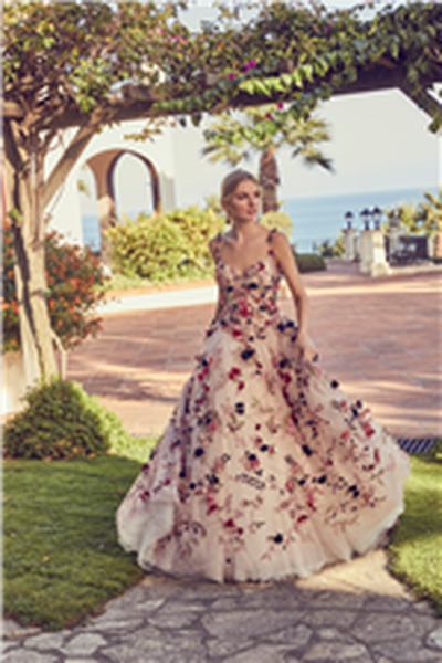 Blonde model stood by a sunny tree near the sea in Ronald Joyce 69426, our Celestina ballgown wedding dress style with multi-coloured flowers and delicate floral applique straps