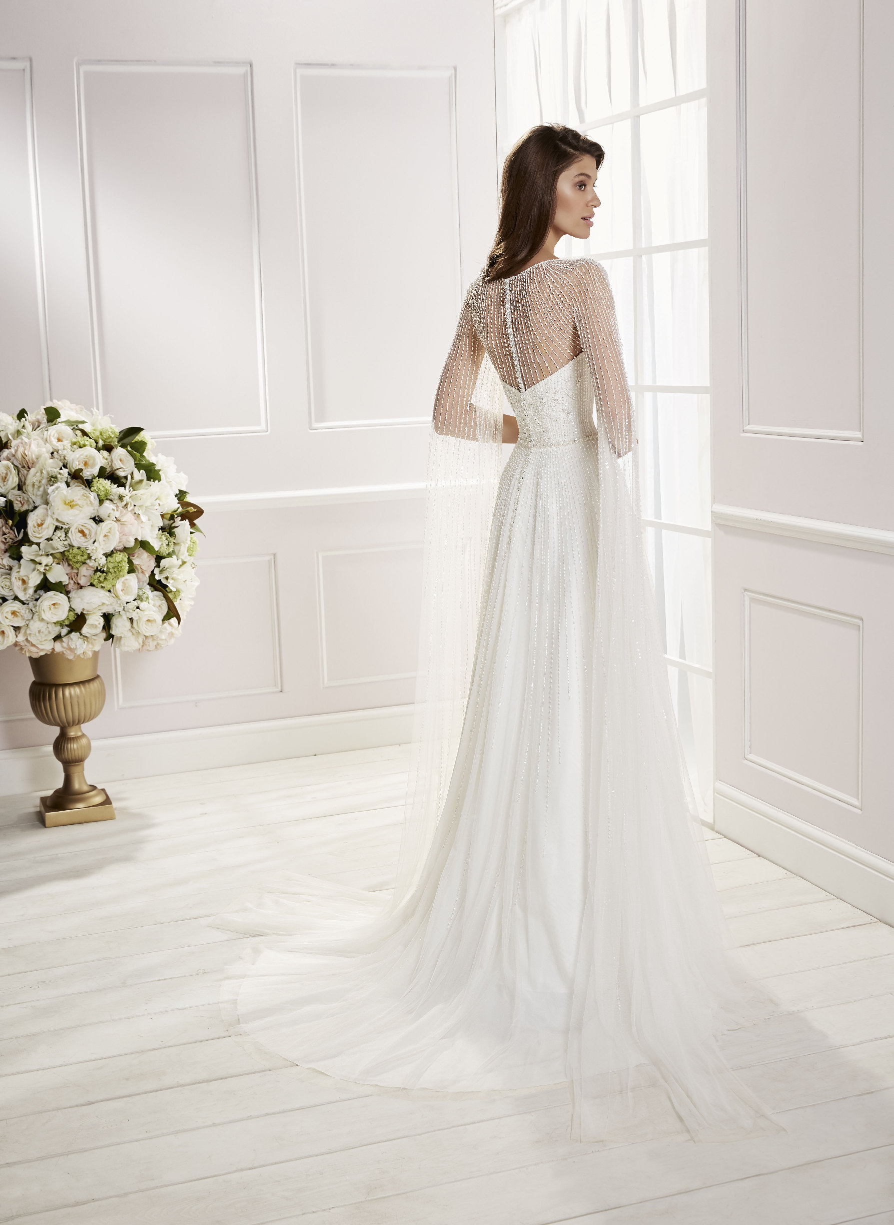 Back image of brunette woman standing in front of a window wearing beaded a-line wedding dress with back bridal button detail