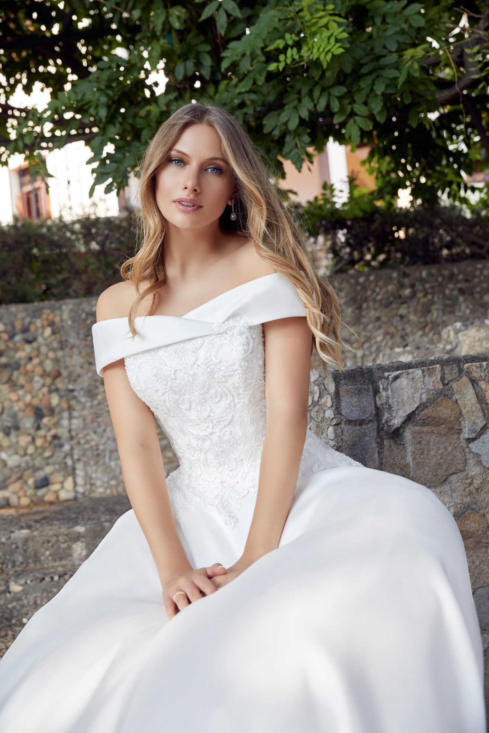 Blonde model sat by a pebbled wall in Ronald Joyce 69501, a plain ivory Mikado ballgown wedding dress with an off the shoulder collar and lace applique bodice