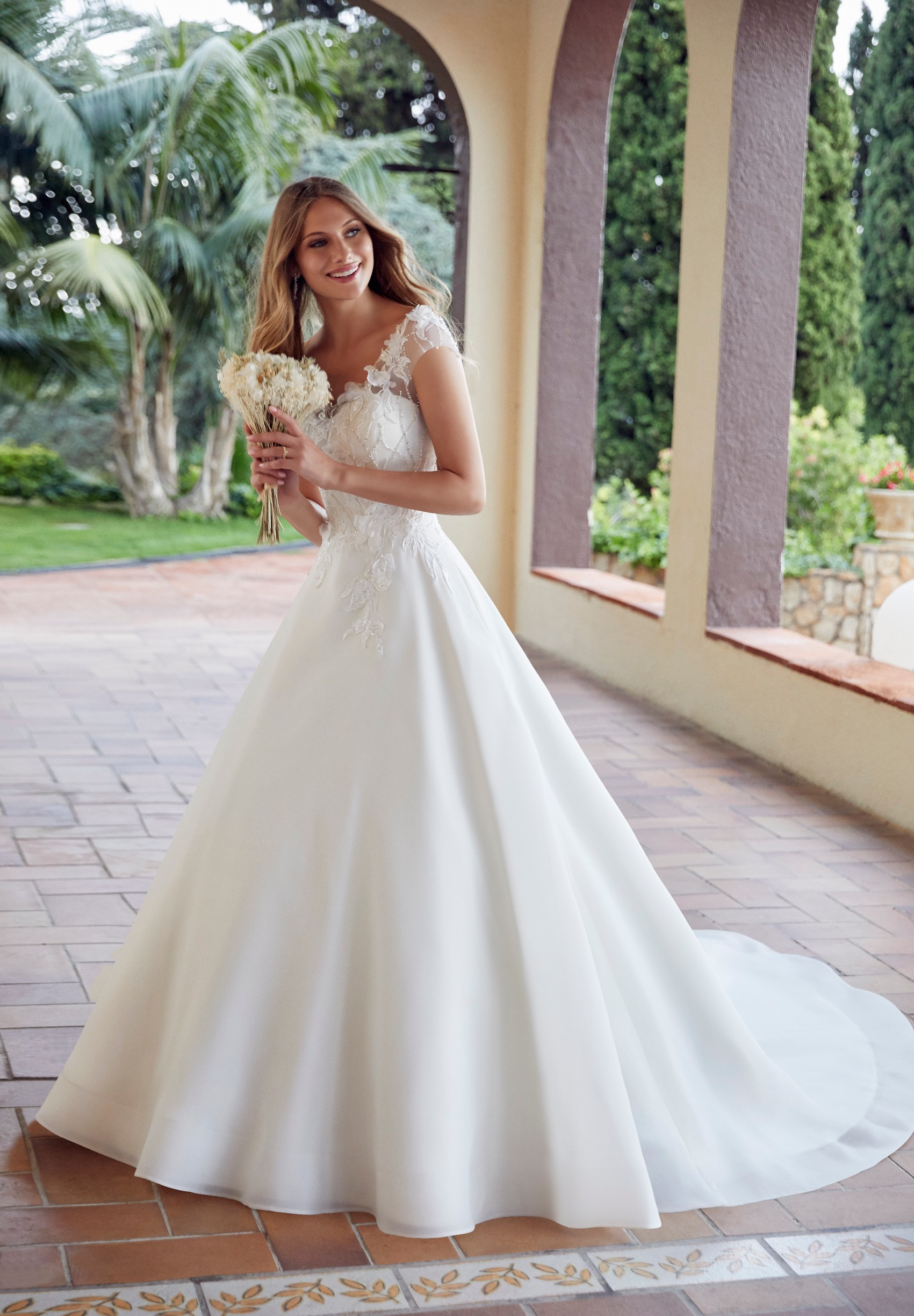 Model stood on a terracotta path by trees in Ronald Joyce 69502, an ivory ballgown wedding dress with a v-neckline, beaded tulle off-the-shoulder cap sleeves and a beaded bodice