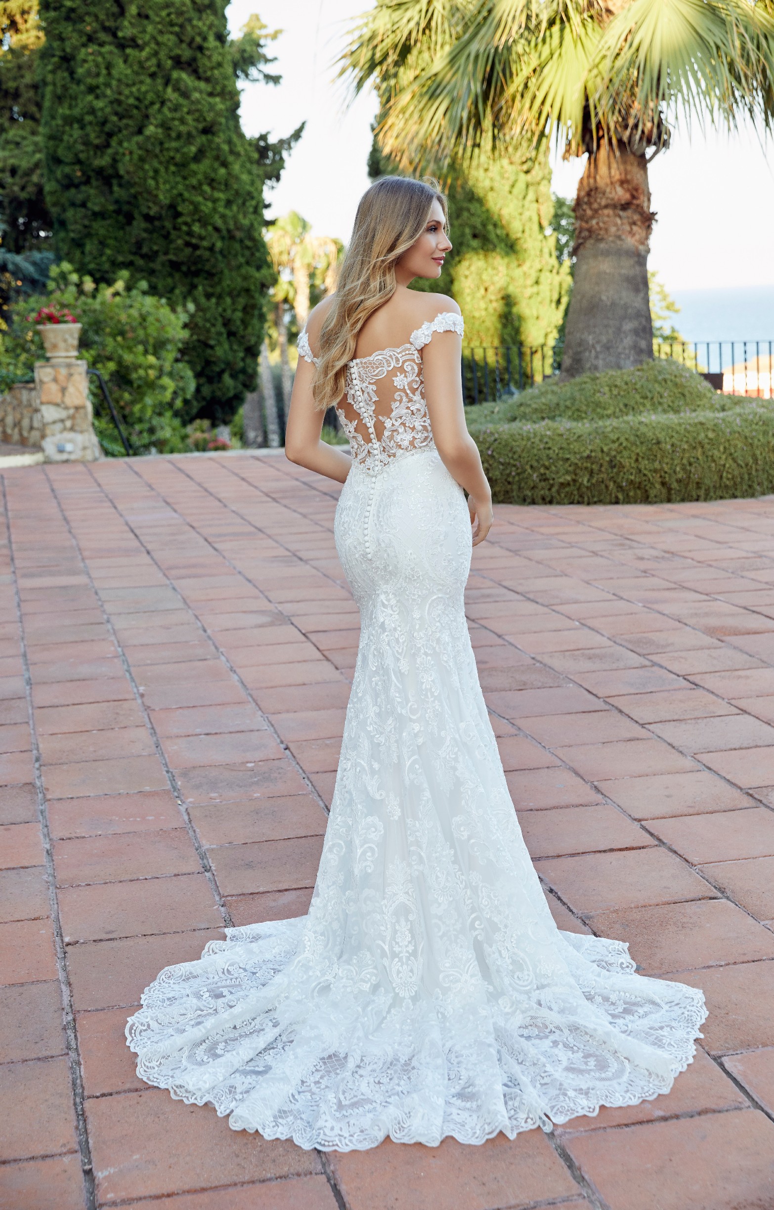Back of model stood on terracotta tiles by a palm tree in Ronald Joyce 69522, a lace fishtail wedding dress with a sweetheart neckline and lace illusion back complete with buttons