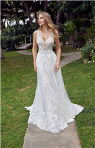 Bride stood by a wood in Ronald Joyce 69525, a lace fit and flare wedding dress with a plunging neckline and bold lace applique skirt hem. Model also wears a detachable A-line overskirt and sparkle belt for two looks.