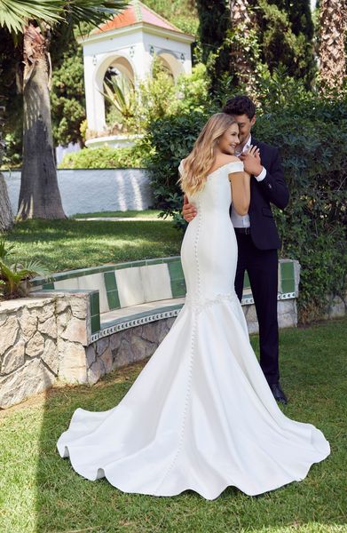 Back of a bride and groom stood in an embrace on a lawn. Bride wears Ronald Joyce 69526, a button backed wedding dress with a plain Mikado fishtail skirt and off the shoulder sleeves