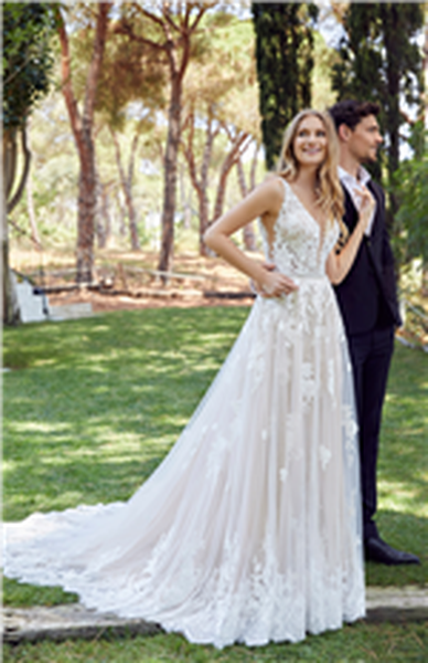 Bride and groom stood on grass by trees. Bride wears Ronald Joyce 69527, a lace, tulle and chiffon fit and flare wedding dress with a plunging illusion neckline and A-line overskirt