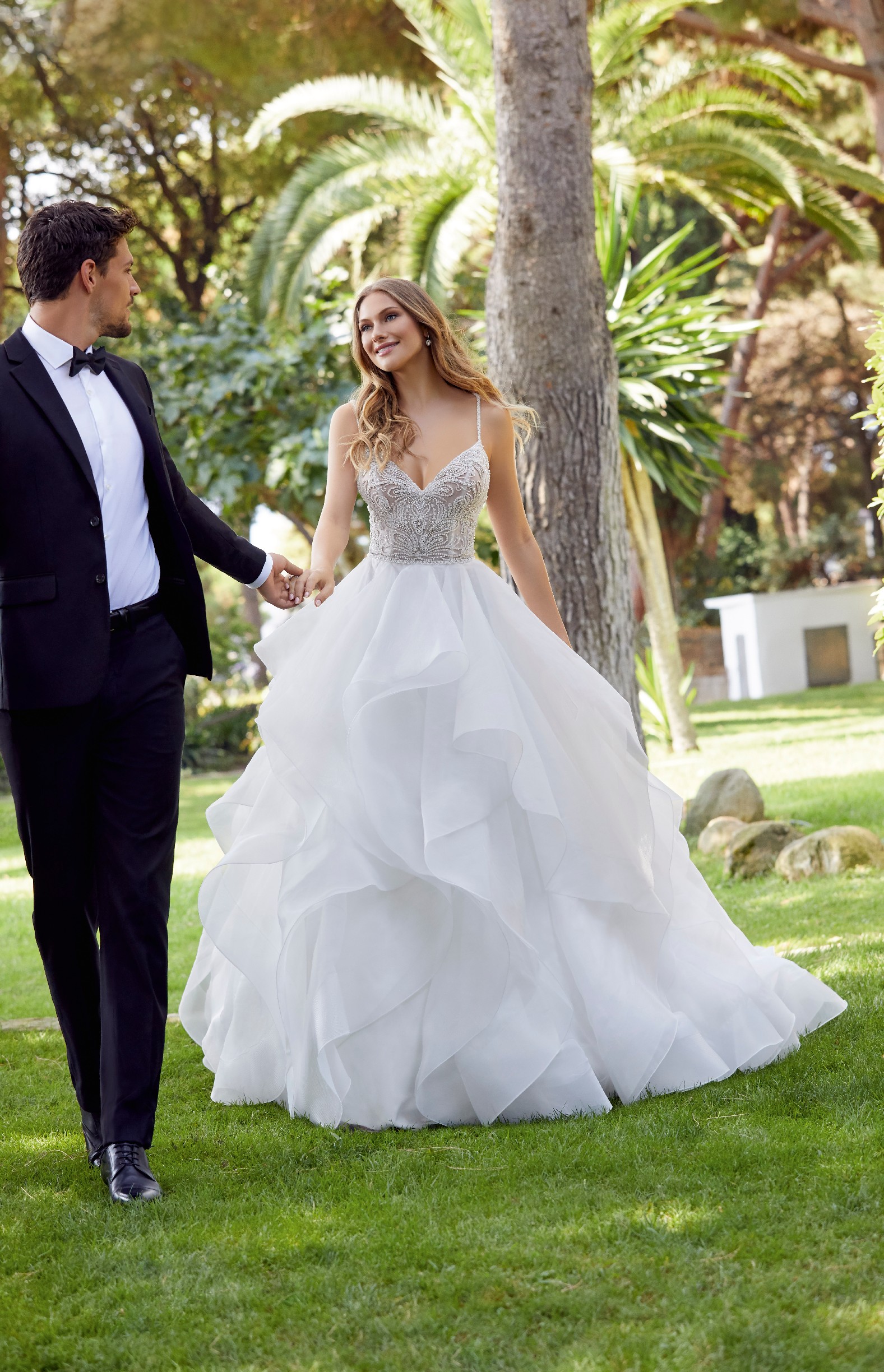 Blonde woman wearing ballgown style wedding dress with beaded bodice and spaghetti straps walking through park with palm trees with man in suit