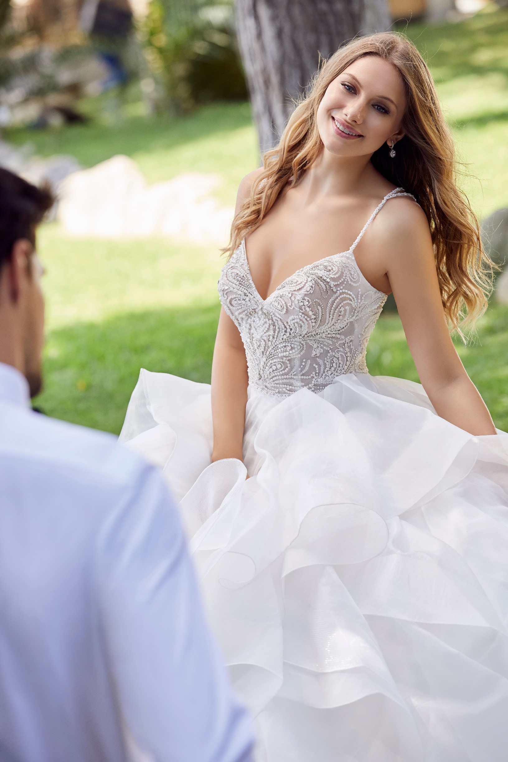 Close up of a model stood by trees opposite a groom in Ronald Joyce 69531, a beaded wedding dress with a pattern beaded bodice, delicate straps and ruffled organza ballgown skirt