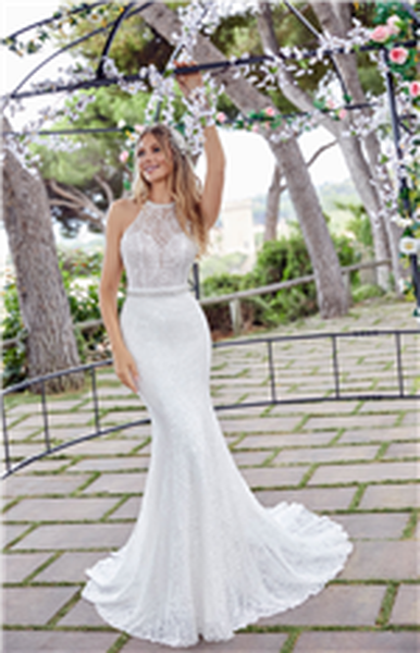 Smiling blonde model stood by trees under a floral decorated metal arbour in Ronald Joyce 69534, a sparkly beaded halter neck wedding dress with a sparkle belt and fishtail skirt