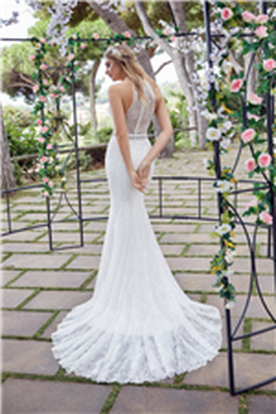 Back image of woman standing under arch decorated with flowers wearing fit and flare wedding dress with beading detail and illusion back