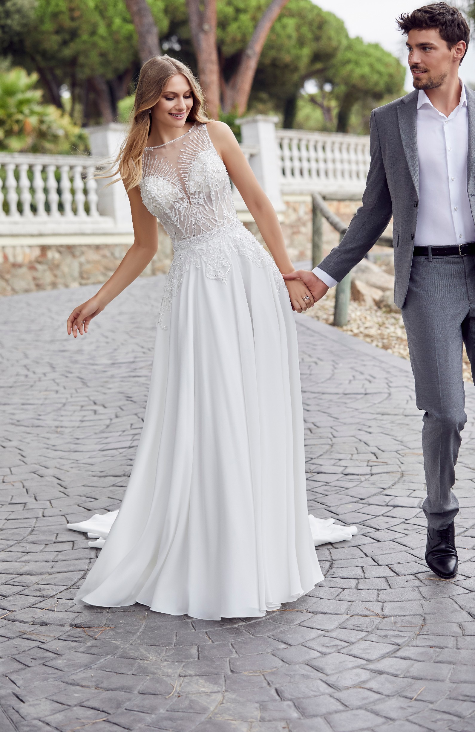 Woman and man walking outside holding hands while the woman wears beaded illusion bodice wedding dress with a-line skirt