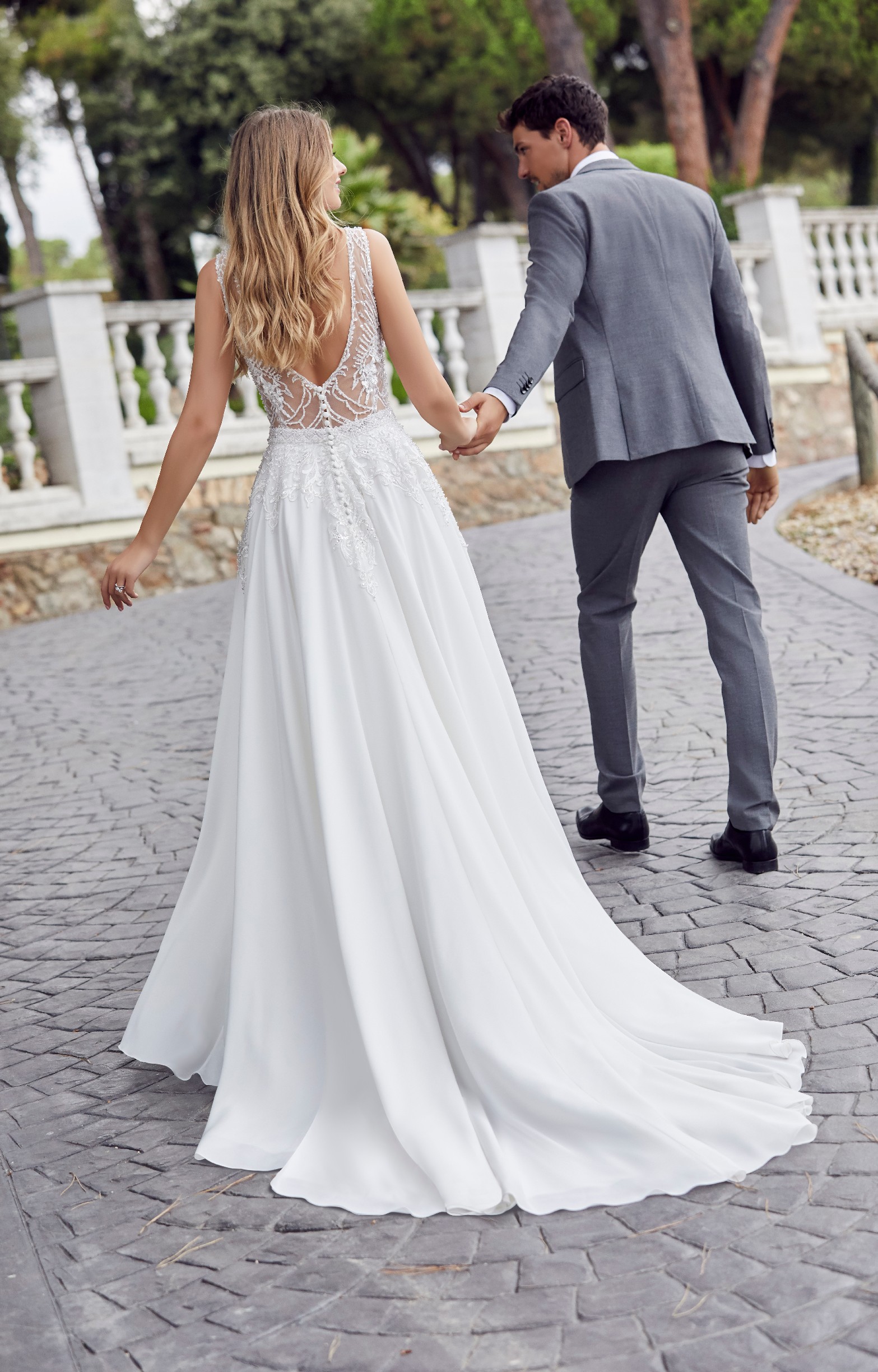 Back image of man and woman wearing a beaded low back wedding dress with contrasting crepe skirt walking hand in hand 