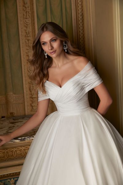 Close up of a model stood in a ballroom in Ronald Joyce 69551, an ivory organdie ballgown wedding dress with a ruched bodice, plain skirt and off the shoulder ruched cap sleeves