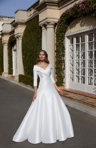 Model stood outside a stately home window in Ronald Joyce 69553, a plain white Mikado ballgown winter wedding dress with an off the shoulder lapel-neckline and ¾ sleeves