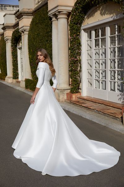 Back of model stood outside a stately home in Ronald Joyce 69553, a plain white Mikado ballgown winter wedding dress with a lapel-neckline, ¾ sleeves and cascading bridal buttons