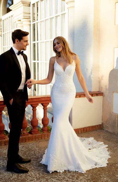 Model stood outside a building next to a groom in Ronald Joyce style 69555, an ivory fit and flare wedding gown with a v-neckline, delicate beaded straps, a lace-beaded body and plain skirt with lace illusion puddle train.