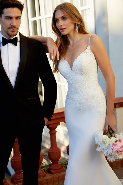 Close up of model stood outside a white building with a groom in Ronald Joyce style 69555, an ivory fit and flare wedding gown with a v-neckline, delicate beaded straps, a lace-beaded body and plain skirt 