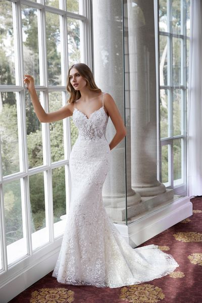 Model stood looking outside floor to ceiling windows in Ronald Joyce style 69560, an ivory fit and flare wedding gown with delicate straps and 3D floral appliques 