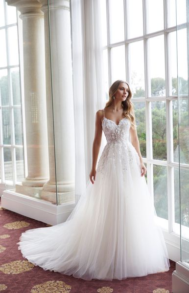 Model stood looking outside grand windows in Ronald Joyce style 69560, a two in one wedding gown with delicate straps, 3D floral appliques, a fit and flare skirt and overskirt