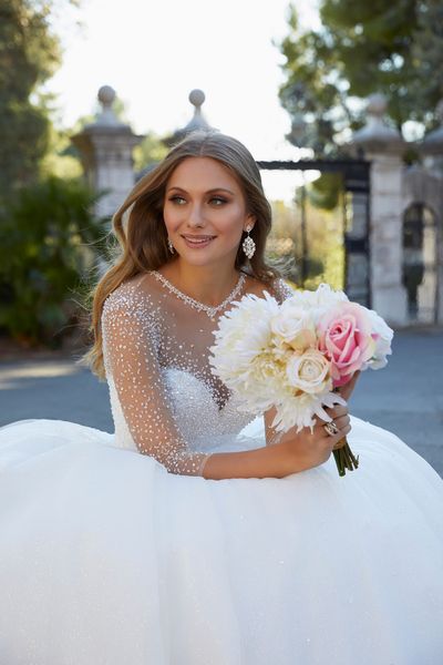 blonde lady sitting down in princess style ballgown wedding dress with beaded bodice and high neckline holding a bouquet of pink and cream flowers