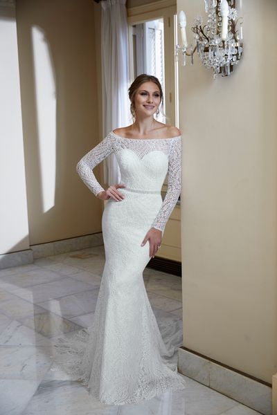 Model stood in a grand room by a wall chandelier in Ronald Joyce 69568, an ivory lace fit and flare wedding dress with a sheer sweetheart neckline and matching long sleeves