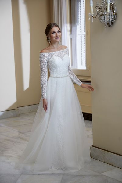 Model stood by a wall chandelier in Ronald Joyce 69568, an ivory lace wedding gown with a sheer sweetheart neckline, matching long sleeves, a fit and flare skirt and overskirt