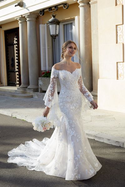 Model stood by a building holding a bouquet in Ronald Joyce 69571, a romantic lace off the shoulder wedding gown with detachable fluted sleeves and an overskirt for two looks