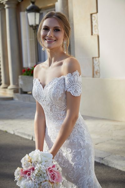Close up of a model stood by a building holding a bouquet in Ronald Joyce 69571, a romantic lace off the shoulder wedding gown with cap sleeves