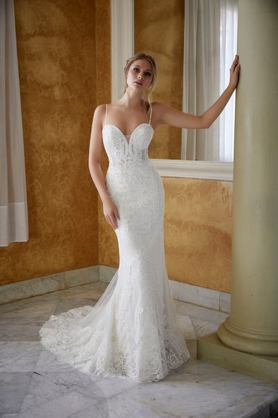 Model stood touching a pillar in an empty room wearing Ronald Joyce 69578, a lace two in one wedding gown with a sweetheart neckline, delicate straps and a fit and flare skirt. 