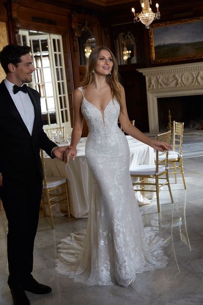 Bride and groom stood in a ballroom near a cream fireplace. She wears Ronald Joyce 695579, a strappy lace fit and flare wedding dress with a plunging neckline and scalloped hem.
