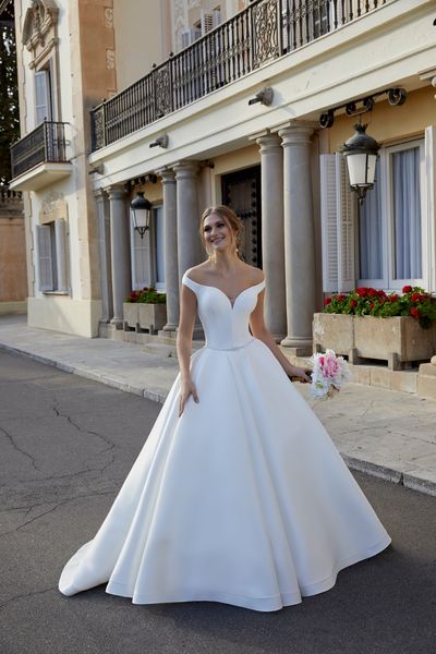 Model stood by a grand building with beautiful red flowerbeds in Ronald Joyce 69583, a plain Mikado off the shoulder ballgown wedding dress with a sweetheart neckline