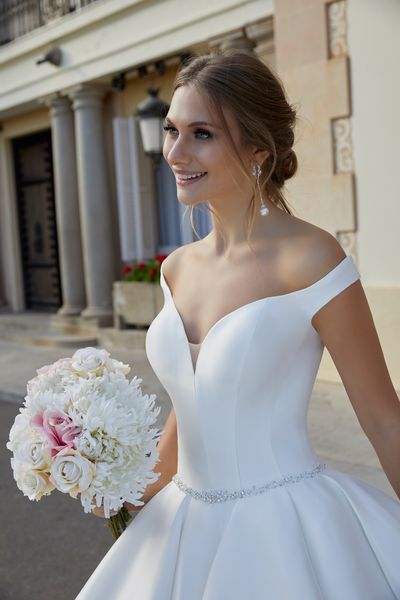 Model stood by a grand building with beautiful red flowerbeds in Ronald Joyce 69583, a plain Mikado off the shoulder ballgown wedding dress with a sweetheart neckline