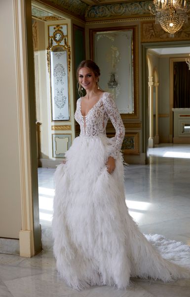 Model stood in a ballroom wearing Ronald Joyce 69584, a glamorous winter wedding dress featuring a lace illusion bodice and matching long sleeves with a feather skirt