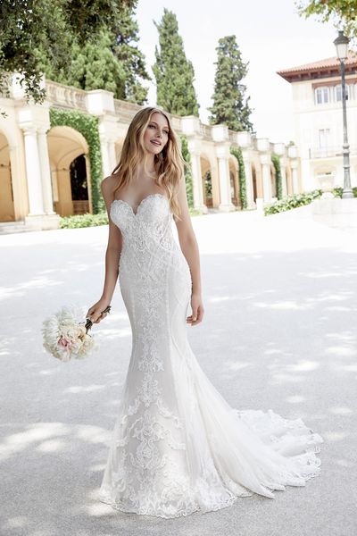 Model stood in a courtyard in Ronald Joyce style 69608, a lace and tulle fit and flare wedding gown with a sweetheart neckline and detachable tulle off-the-shoulder short sleeves 