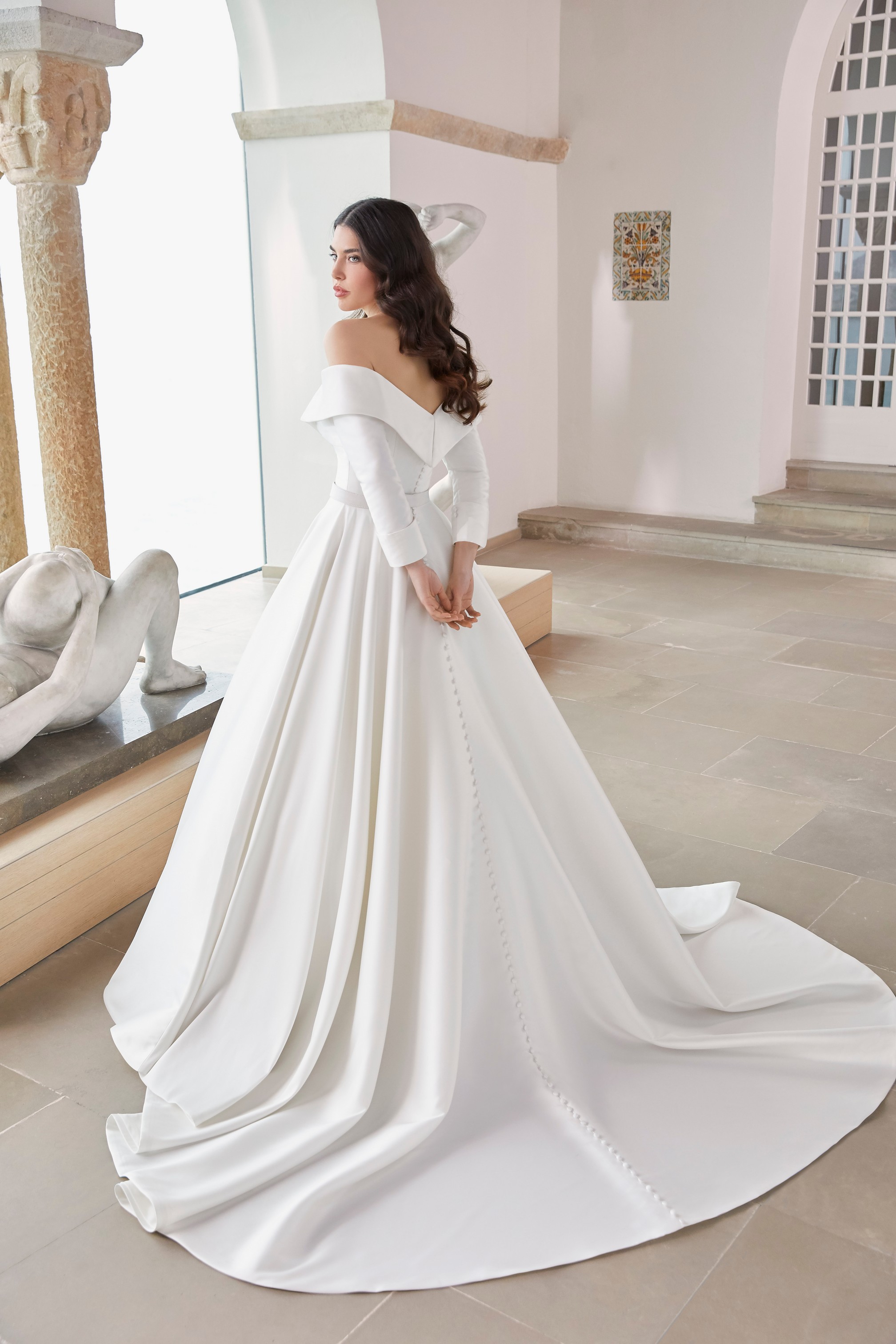 Back image of a brunette woman standing in museum in mikado ballgown wedding dress with v neck back with bridal buttons
