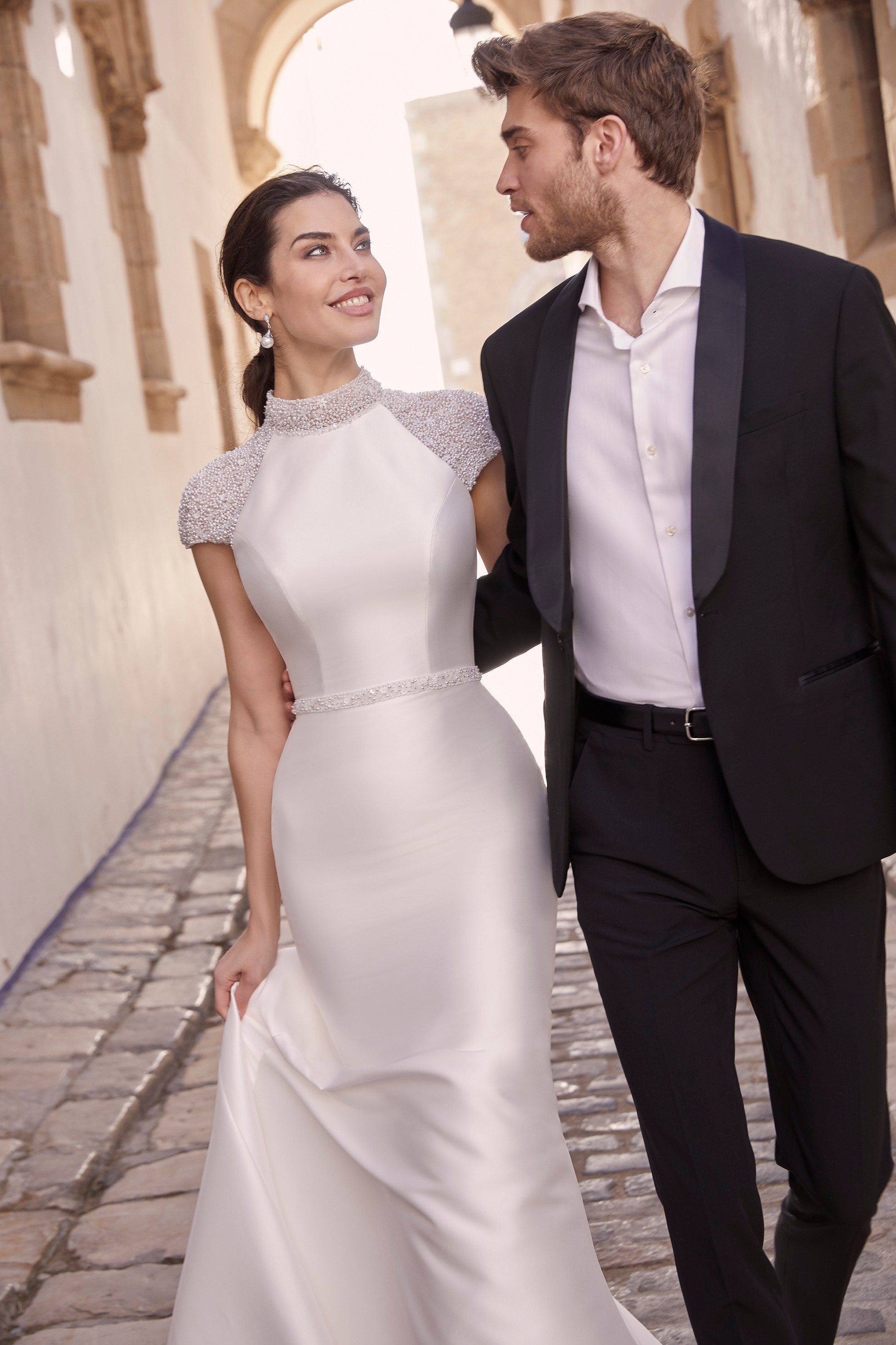 Man and woman walking down quaint backstreet while woman wears mikado fit and flare wedding dress with beaded cap sleeves and embellished waist belt