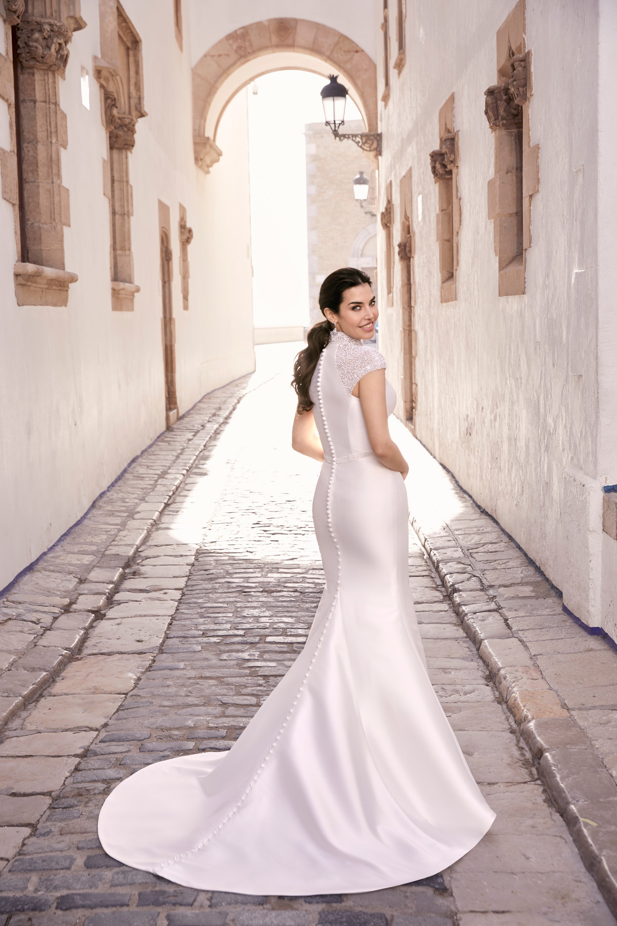 Woman stood in idyllic European side street wearing fitted mikado wedding dress with bridal button detail and embellished cap sleeves