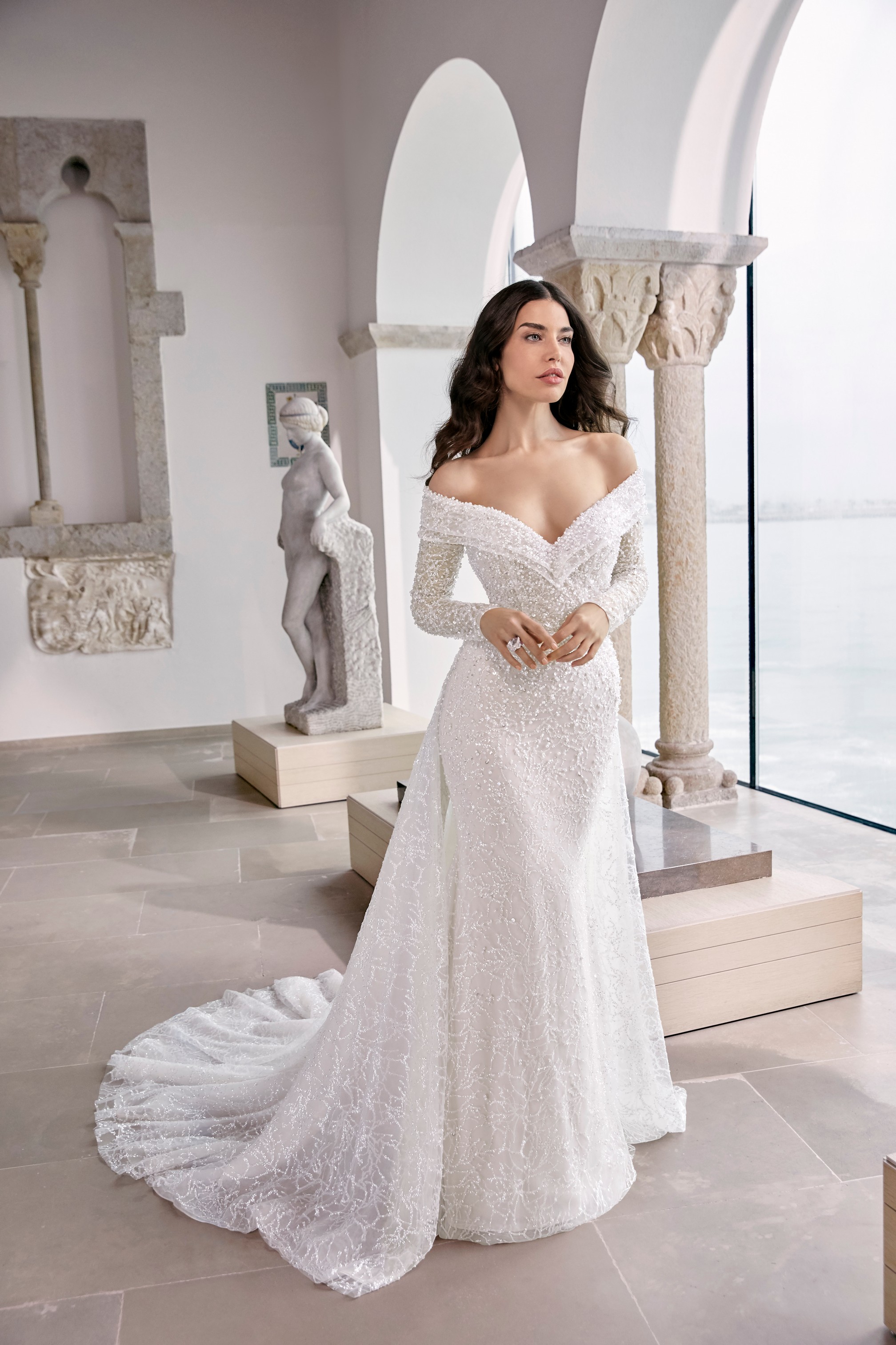 Model stood by statues in Ronald Joyce 69659, a heavily beaded wedding dress with an off the shoulder v-neckline, collar and detachable long sleeves. Model also wears overskirt