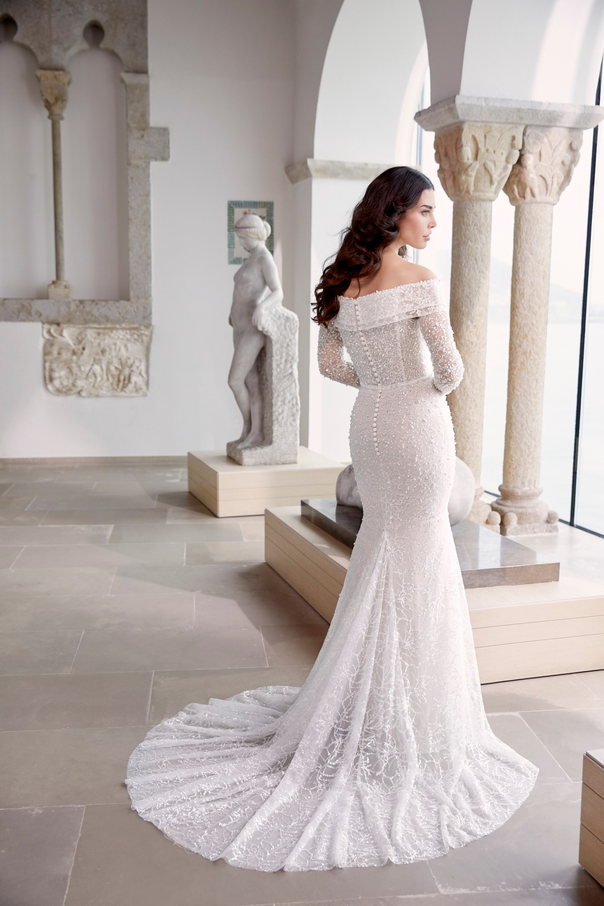 Back of model stood by statues in Ronald Joyce 69659, an exquisitely beaded wedding dress with an off the shoulder v-neckline, lapel-style collar and detachable long sleeves