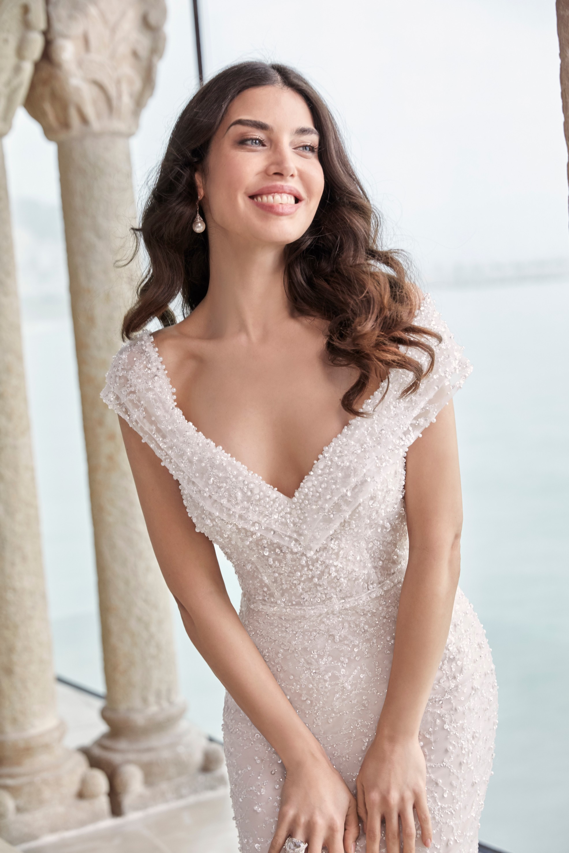 Brunette lady standing in museum in front of historical pillars and overlooking the ocean wearing heavily beaded fit and flare wedding dress with off the shoulder detail and pearl drop earrings
