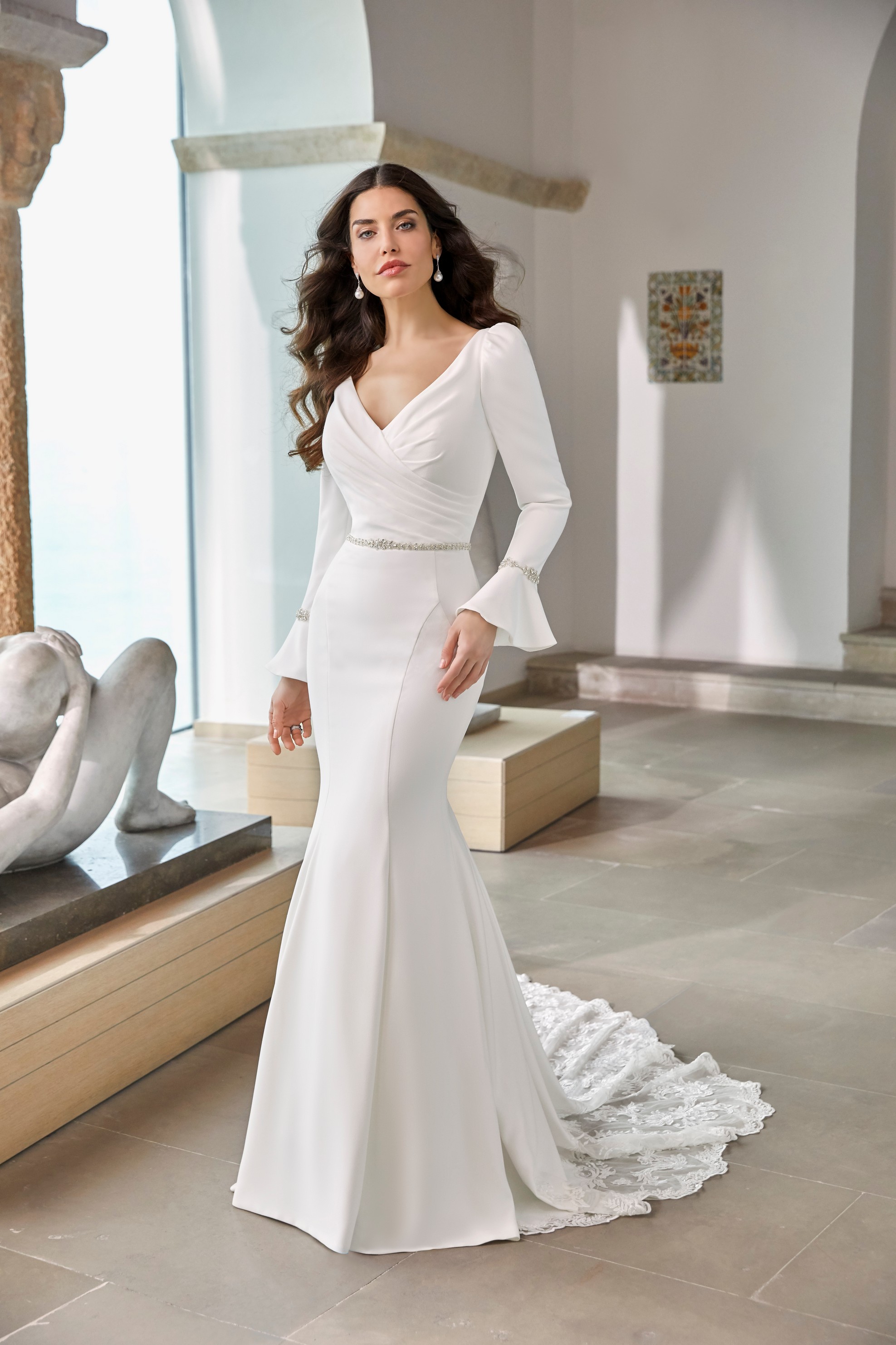 Model stood in an empty museum room in Ronald Joyce 69668, a long sleeved plain wedding dress with fluted cuffs, a sparkle belt, v-neckline and fit and flare skirt with lace train