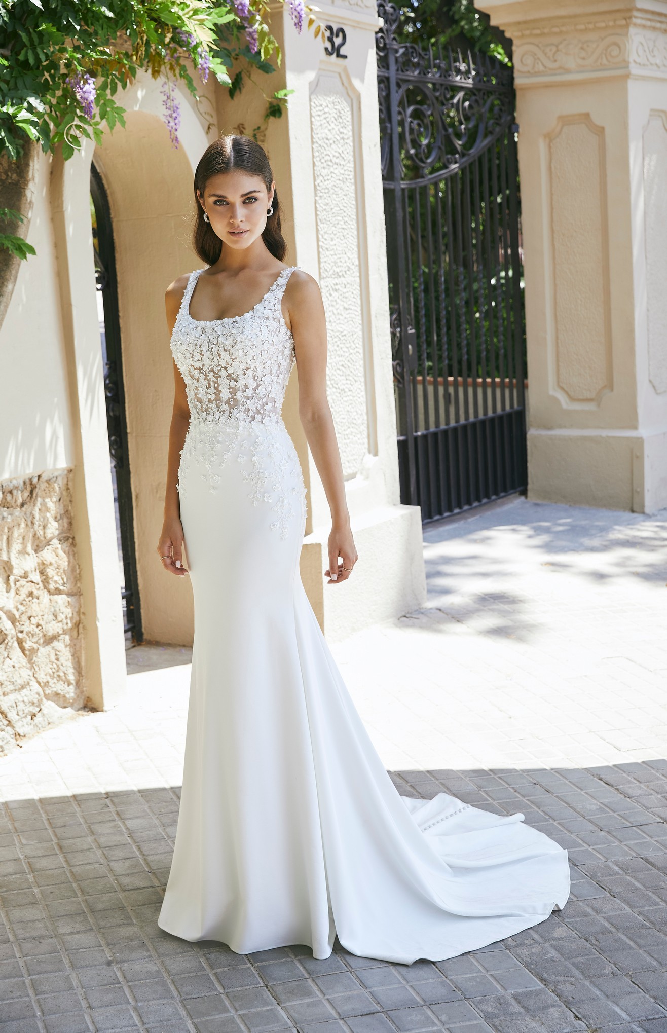 Model by a sunny black gate in Ronald Joyce 69702, a fit and flare wedding dress with a feminine scoop neckline, 3D floral appliqué bodice and plain skirt.