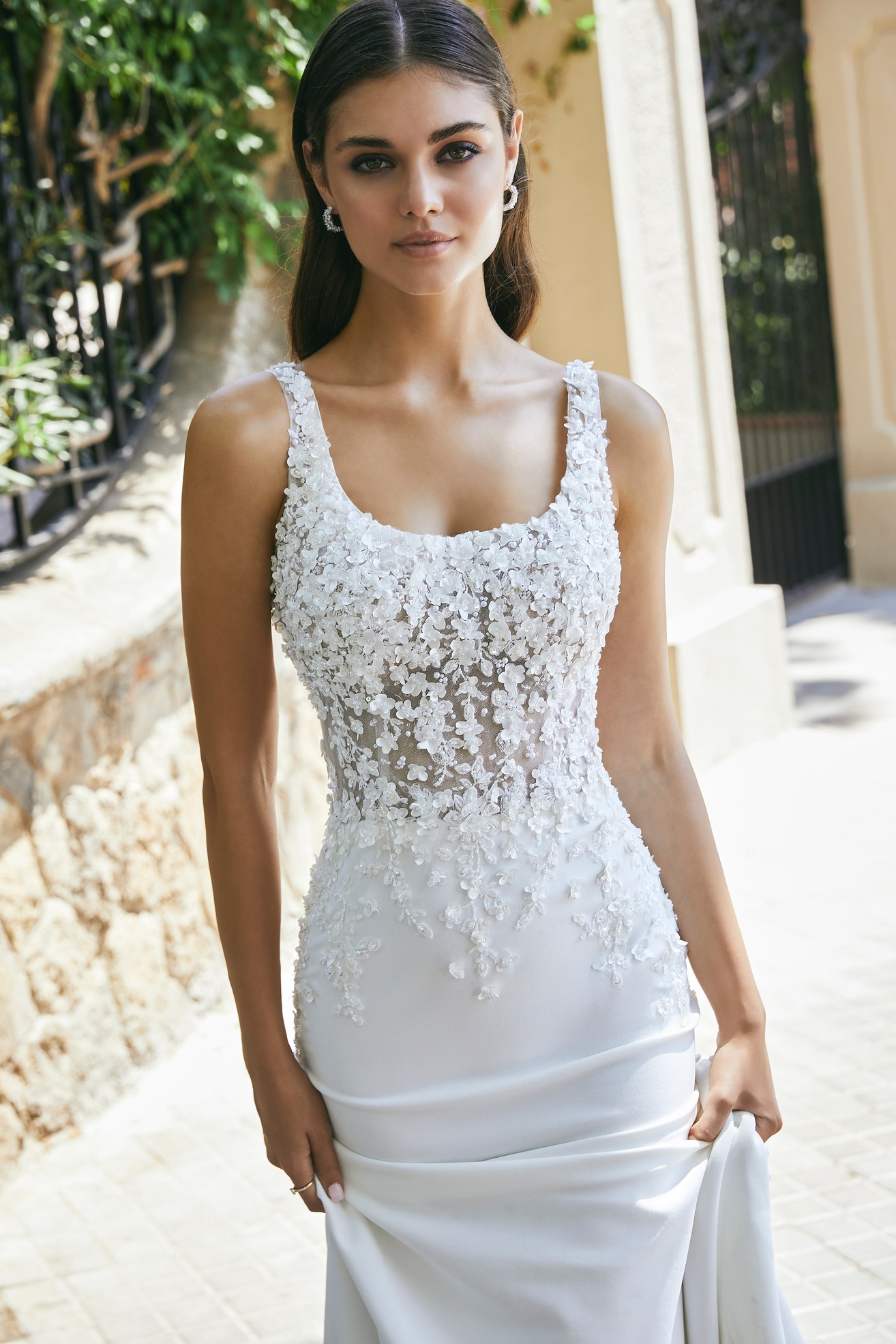 Close up of model stood outside by a sunny gate in Ronald Joyce 69702, a fit and flare wedding dress with a scoop neckline, 3D floral bodice and plain skirt.