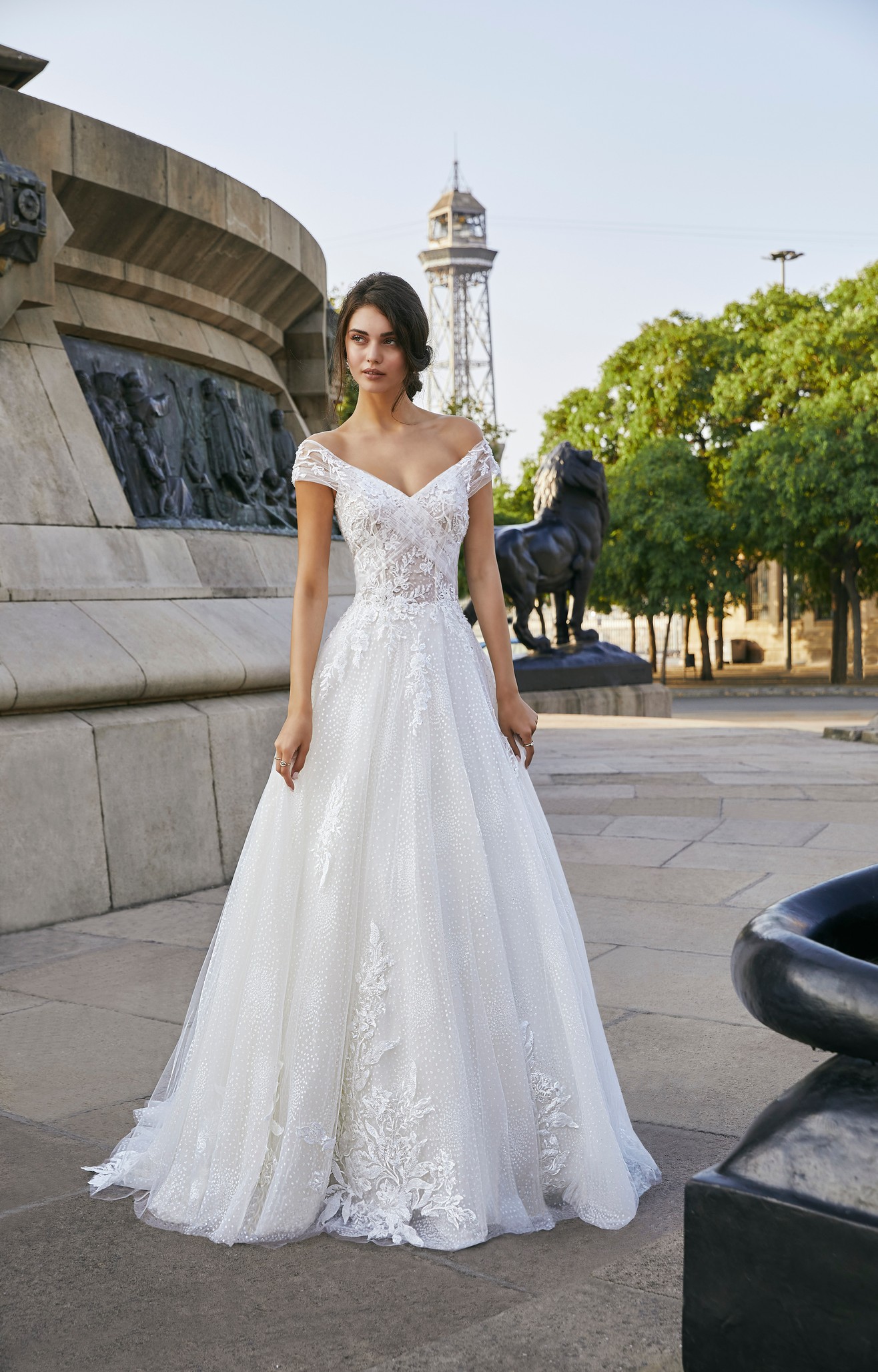 Model stood outside in Ronald Joyce style 69703, an organza A-line wedding dress with off-the-shoulder capped sleeves and a lace applique tulle skirt