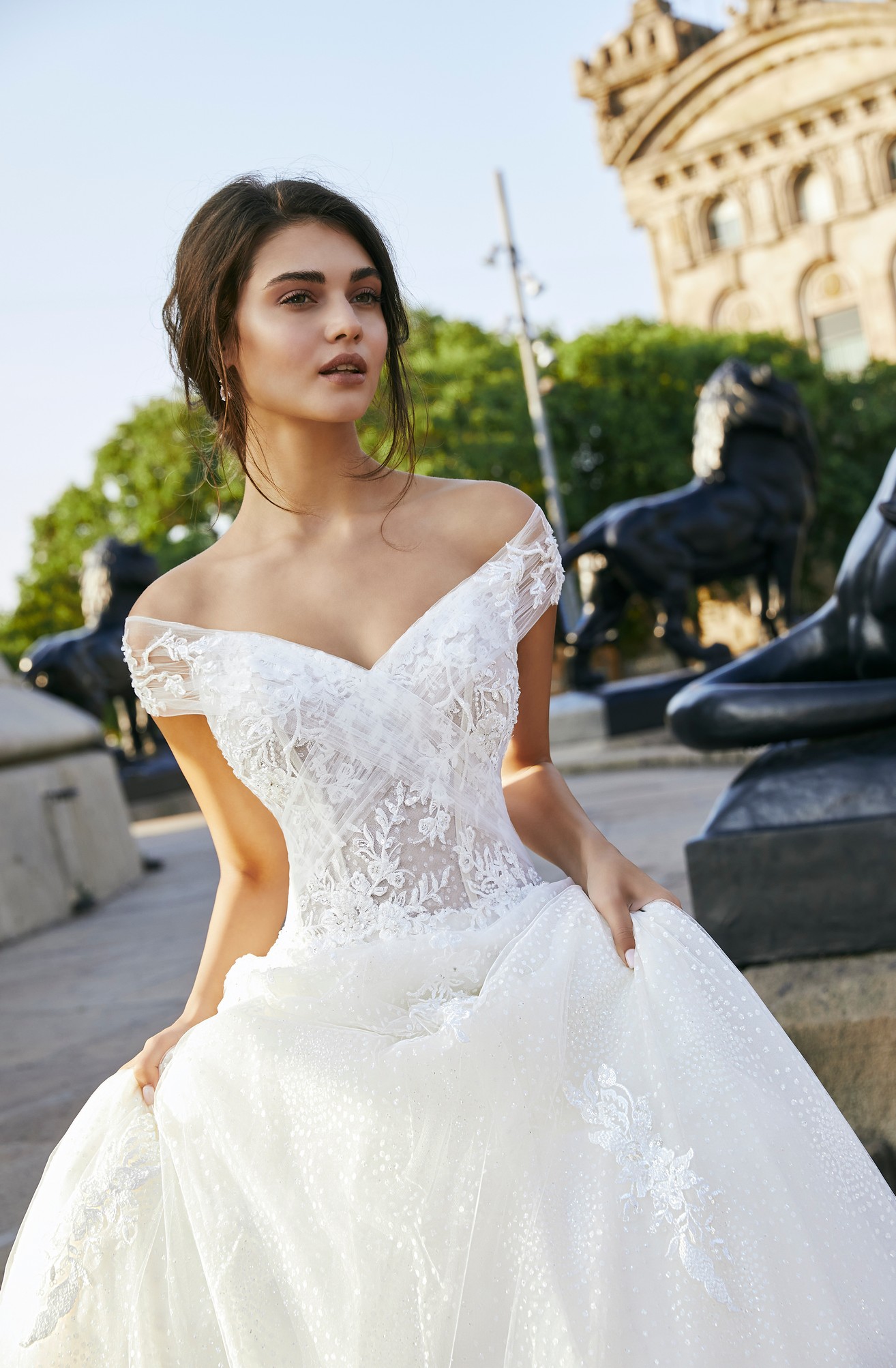 Close up of a model stood by lion statues in Ronald Joyce 69703, an off-the-shoulder A-line wedding dress with cap sleeves, an illusion bodice and v-neckline in tulle and lace applique bridal fabrics